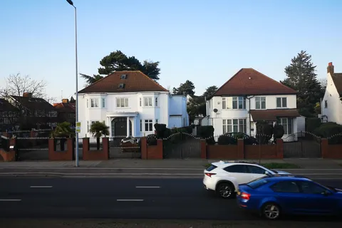 White-rendered interwar houses with hipped tiled roofs, a two-storey side addition and roof windows, behind brick walls on an outer London main road
