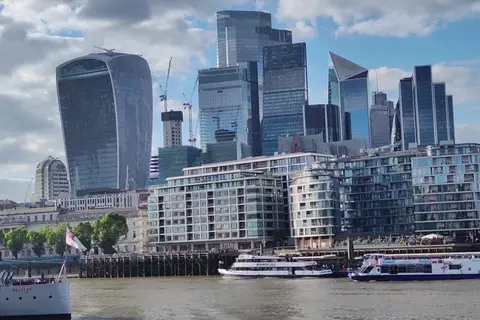Glass-fronted blocks of flats on the north bank of the Thames, with the City of London's office towers, including the Walkie-Talkie, rising behind and river boats passing in front