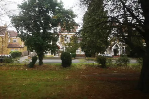 Large yellow-brick Victorian and Edwardian houses with gables and a timbered porch, seen through trees across a grassy green on an overcast autumn day