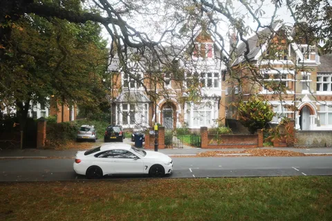 Large red-brick Victorian-style houses with white gables and bay windows across a road from a grassy green, with a white car passing and autumn leaves on the verge