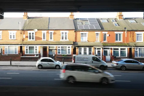 A row of red-brick terraced houses seen from beneath a road flyover, with roof windows and solar panels in the front roof slopes and cars passing on the main road in front