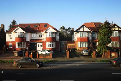 Two pairs of interwar semi-detached houses with red-tiled roofs, bay windows and paved front drives beside a main road, the left-hand pair with roof windows in the front roof slope and the right-hand pair with mock-Tudor gables